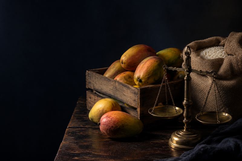A wooden crate of ripe mangoes and a sack of rice beside an antique brass balance scale on dark wood