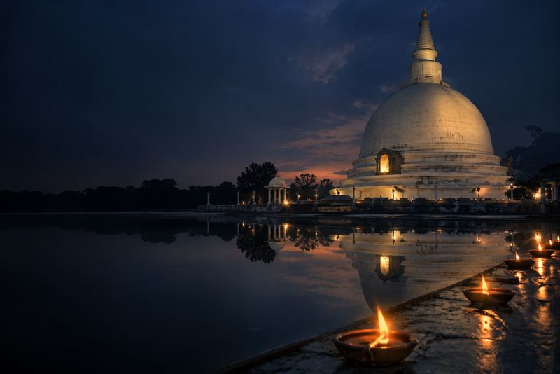 A vast white stupa at dusk reflected in still water, a few oil lamps glowing, dark blue sky