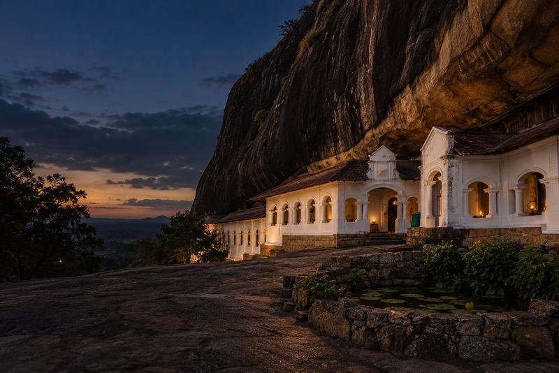 The Dambulla cave temple in Sri Lanka at dusk: a long whitewashed colonnaded front with arched doorways and red clay-tiled roofs, built under the overhang of a huge dark granite rock, oil lamps glowing in the doorways, no people. Real Sri Lankan Buddhist architecture, NOT Indian or Petra-style carved rock facades