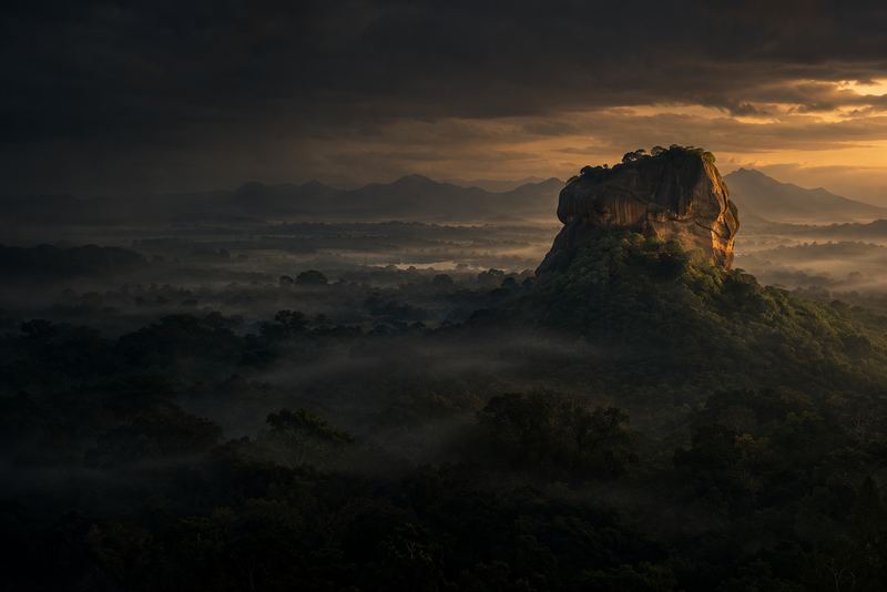 Sigiriya rock rising from misty forest at dawn, seen from a distance, dark moody sky with a gold light on the rock's face