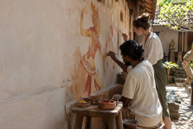 A muralist and a visitor painting on lime plaster in a courtyard