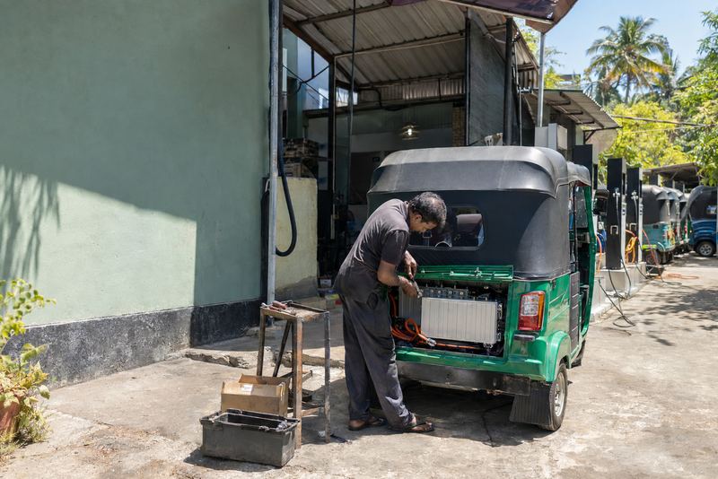 A mechanic fitting a battery into a three-wheeler at a sunny garage