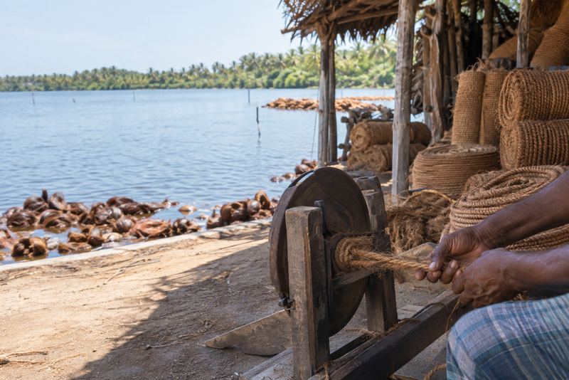 Hands twisting coir rope on a wheel beside a lagoon