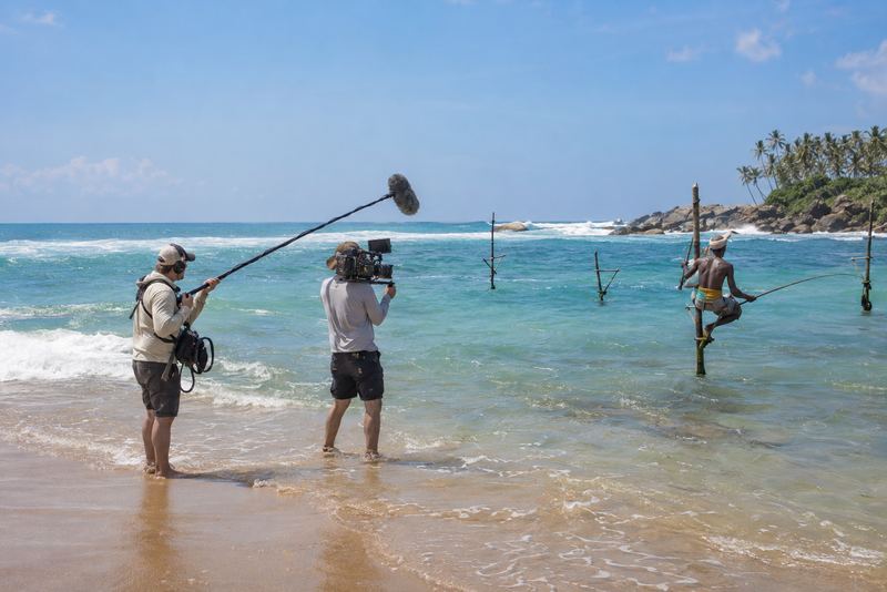 A camera crew filming a stilt fisherman in bright sunshine