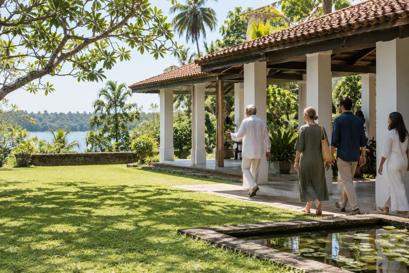 Visitors touring a sunlit Bawa garden pavilion by a lake